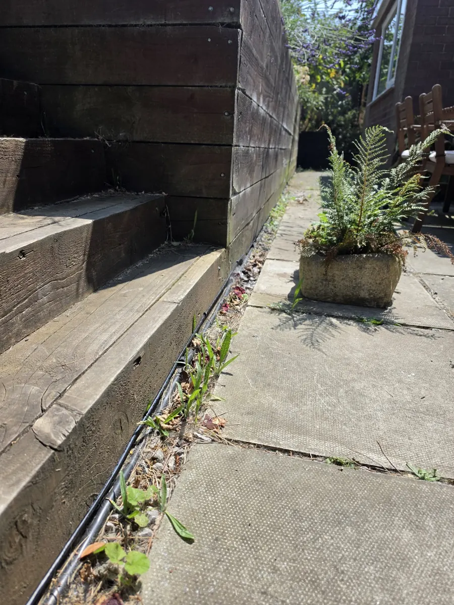 Black external data cable clipped along the base of a timber sleeper wall beside paving